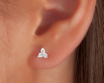 May include: A small, silver stud earring featuring three clear gemstones in a triangular arrangement. The earring is shown in situ on an ear, showcasing its subtle sparkle and elegant design. The gemstones are round-cut.