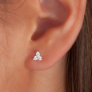 Op de afbeelding: Kleine, zilveren oorbel met drie heldere, rond geslepen edelstenen, gerangschikt in een driehoekige cluster. De oorbel wordt op een oor gedragen, waardoor het delicate ontwerp en de glans worden benadrukt.