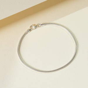 May include: A silver snake chain bracelet with a lobster clasp. The bracelet is made of a flexible, rounded chain and is displayed on a cream-coloured surface. A delicate piece of jewellery.