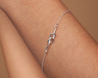 May include: A silver bracelet featuring a knot design. The bracelet is made of a delicate chain with a knot-shaped centerpiece. The bracelet is shown on a person's arm against a neutral background.