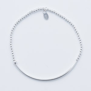 Może przedstawiać: Delikatna srebrna bransoletka z małymi, okrągłymi koralikami i zakrzywionym prętem. Bransoletka ma minimalistyczny design i zawiera małą zawieszkę z napisem "925". Biżuteria jest prezentowana na białym tle.