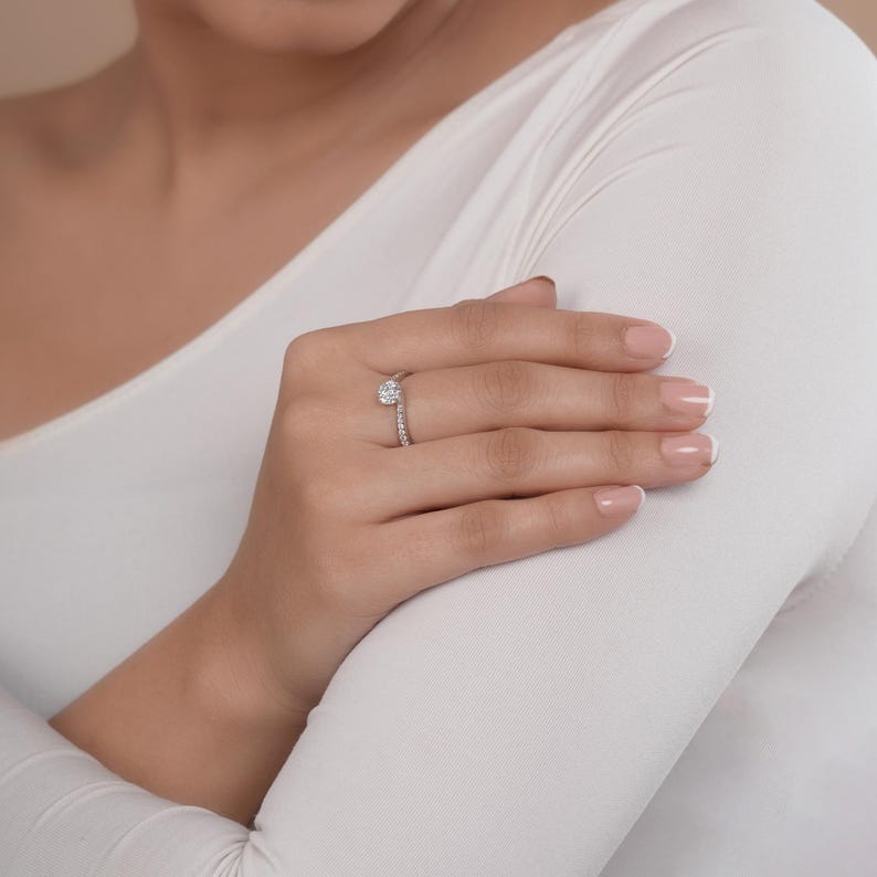 May include: A silver engagement ring featuring a round, clear central stone and a band set with smaller stones. The ring is worn on a finger, with a white long-sleeved top visible in the background.