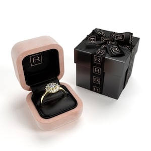 May include: A gold ring with a large, round, clear gemstone surrounded by smaller stones, displayed in an open pink velvet ring box with a black interior. A black gift box with a black ribbon is next to the ring box.