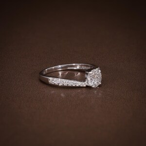 May include: A silver engagement ring with a large, round, clear diamond centre stone. The band features smaller accent stones set along one side. The ring is displayed on a brown surface, highlighting its sparkle and design.