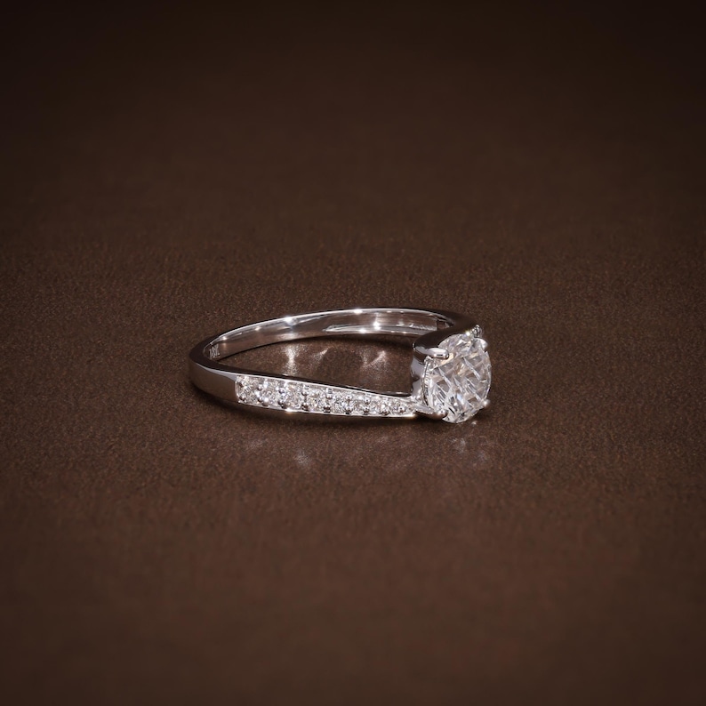 May include: A silver engagement ring with a large, round, clear diamond centre stone. The band features smaller accent stones set along one side. The ring is displayed on a brown surface, highlighting its sparkle and design.