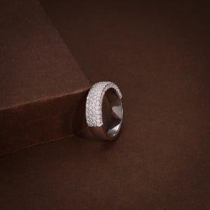 May include: A silver ring with a band covered in small, sparkling white stones. The ring is set against a brown background, creating a contrast that highlights the ring's brilliance. The ring is a piece of jewelry.