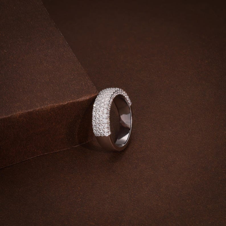 May include: A silver ring with a band covered in small, sparkling white stones. The ring is set against a brown background, creating a contrast that highlights the ring's brilliance. The ring is a piece of jewelry.