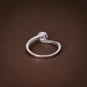May include: A silver ring featuring a central, clear gemstone. The band has a flowing design, with smaller stones set along one side. The ring is displayed on a brown surface, accentuating its brilliance.