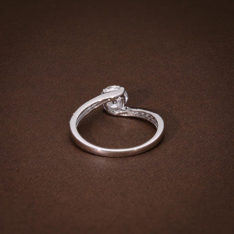 May include: A silver ring featuring a central, clear gemstone. The band has a flowing design, with smaller stones set along one side. The ring is displayed on a brown surface, accentuating its brilliance.
