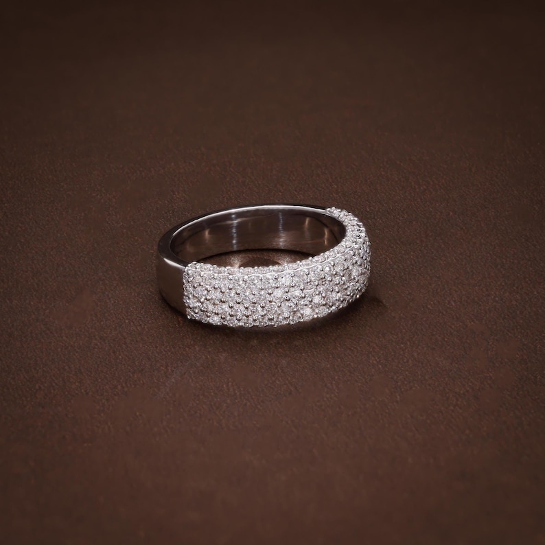 May include: A silver ring with a band encrusted with small, clear gemstones. The ring is set against a brown background, showcasing its sparkling details. The gemstones cover the top and sides of the ring.
