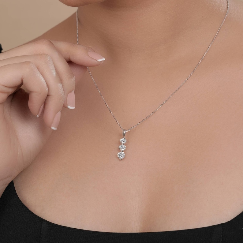 May include: A delicate silver necklace featuring three round, sparkling gemstones in a vertical arrangement. The pendant hangs from a fine chain, creating a simple yet elegant design. The necklace is shown against a dark background.