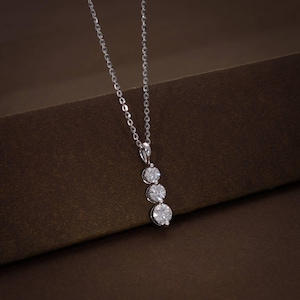 May include: A silver necklace with three round, sparkling gemstones in a vertical line. The pendant hangs from a delicate chain, set against a brown background. The gemstones appear to be diamonds.