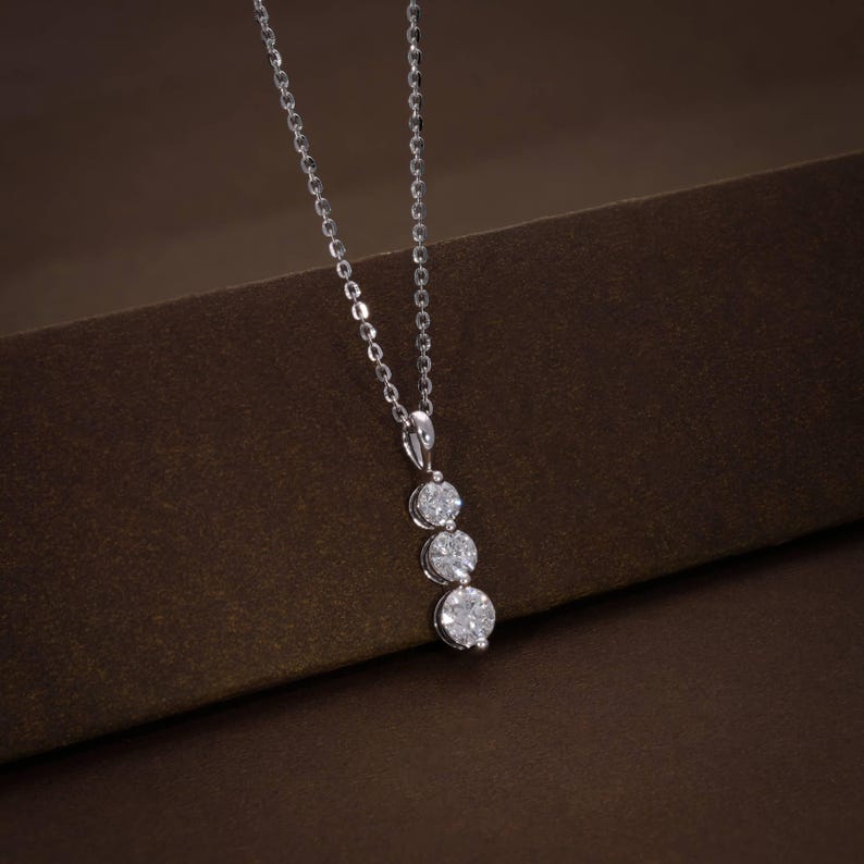 May include: A silver necklace with three round, sparkling gemstones in a vertical line. The pendant hangs from a delicate chain, set against a brown background. The gemstones appear to be diamonds.