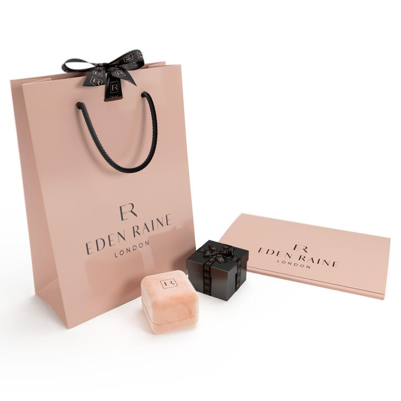 May include: A blush pink shopping bag, ring box, and gift box with black ribbon. The bag and card feature the text "EDEN RAINE LONDON". The ring box is a light peach color.