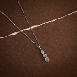May include: A delicate silver necklace featuring three round, sparkling gemstones. The pendant hangs from a fine chain, set against a brown background with a white, torn paper accent. The necklace is a classic piece of jewelry.
