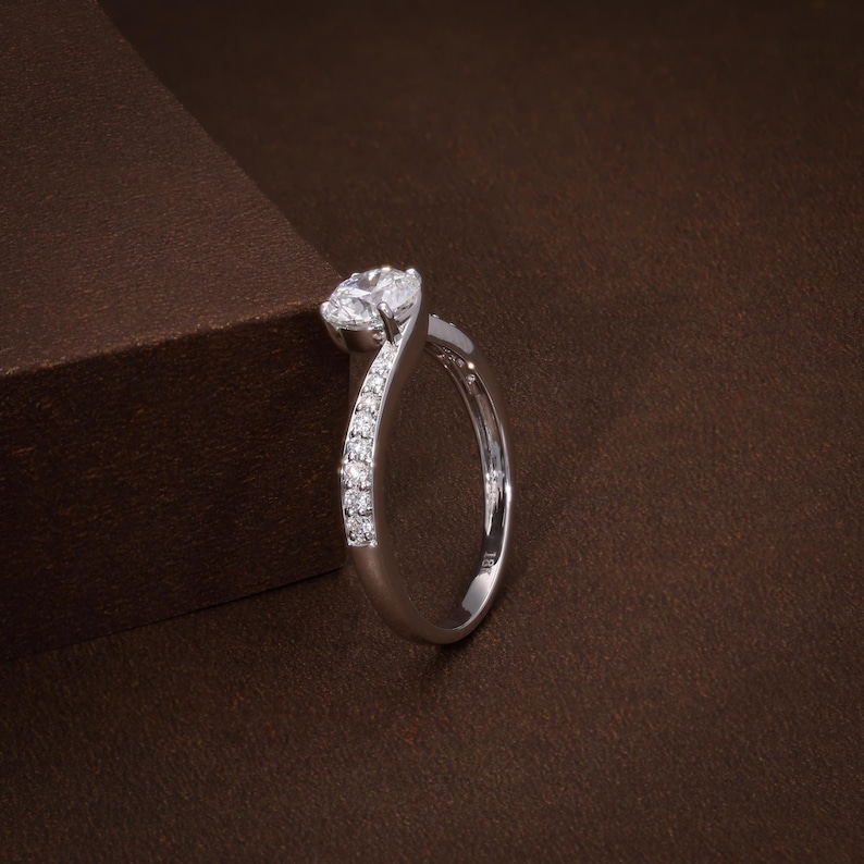 May include: A silver ring featuring a large, round, clear gemstone. The band curves elegantly, with smaller gemstones set along one side. The ring is presented against a brown backdrop.
