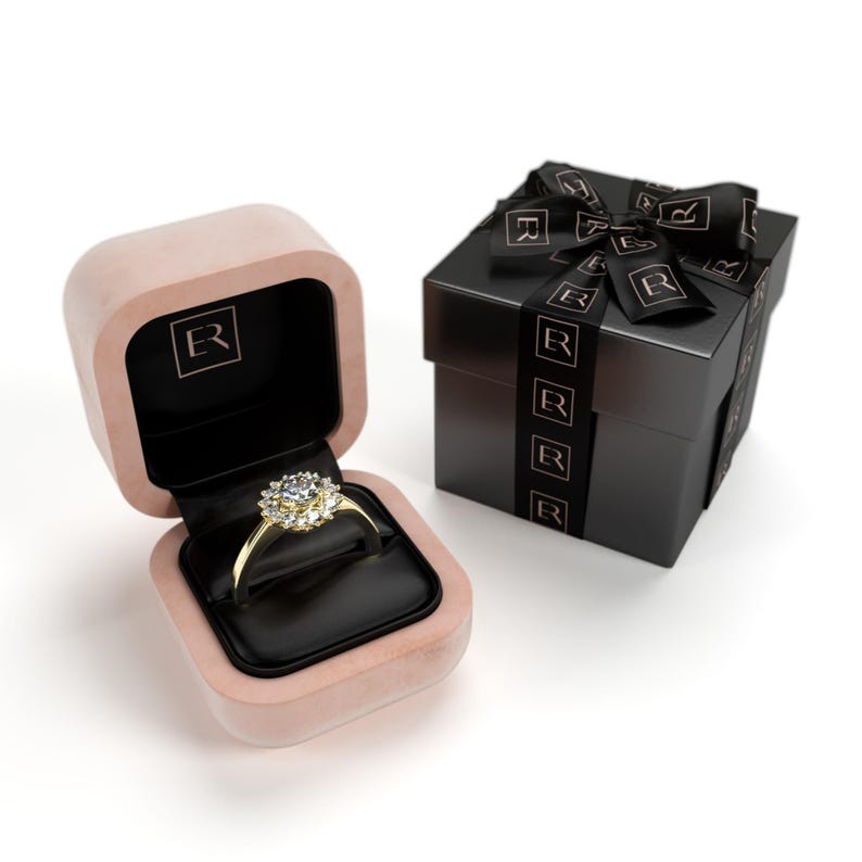 May include: A gold ring with a large, round, clear gemstone surrounded by smaller stones, displayed in an open pink velvet ring box with a black interior. A black gift box with a black ribbon is next to the ring box.