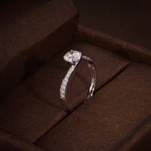 May include: A silver ring featuring a large, clear, round-cut diamond. The band curves with smaller diamonds set along one side. The ring is presented in a brown jewellery box. The ring is marked with '18K'.
