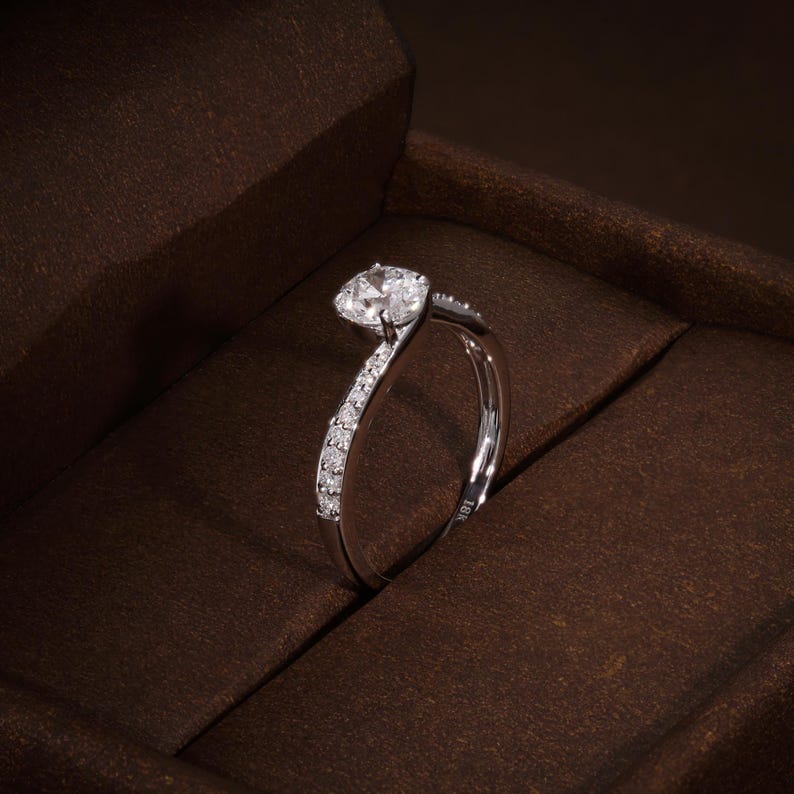 May include: A silver ring featuring a large, clear, round-cut diamond. The band curves with smaller diamonds set along one side. The ring is presented in a brown jewellery box. The ring is marked with '18K'.