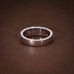 May include: A silver ring with a band of small, clear gemstones set around the top edge. The ring is set against a brown background, showcasing its polished metal and sparkling stones. A classic piece of jewelry.