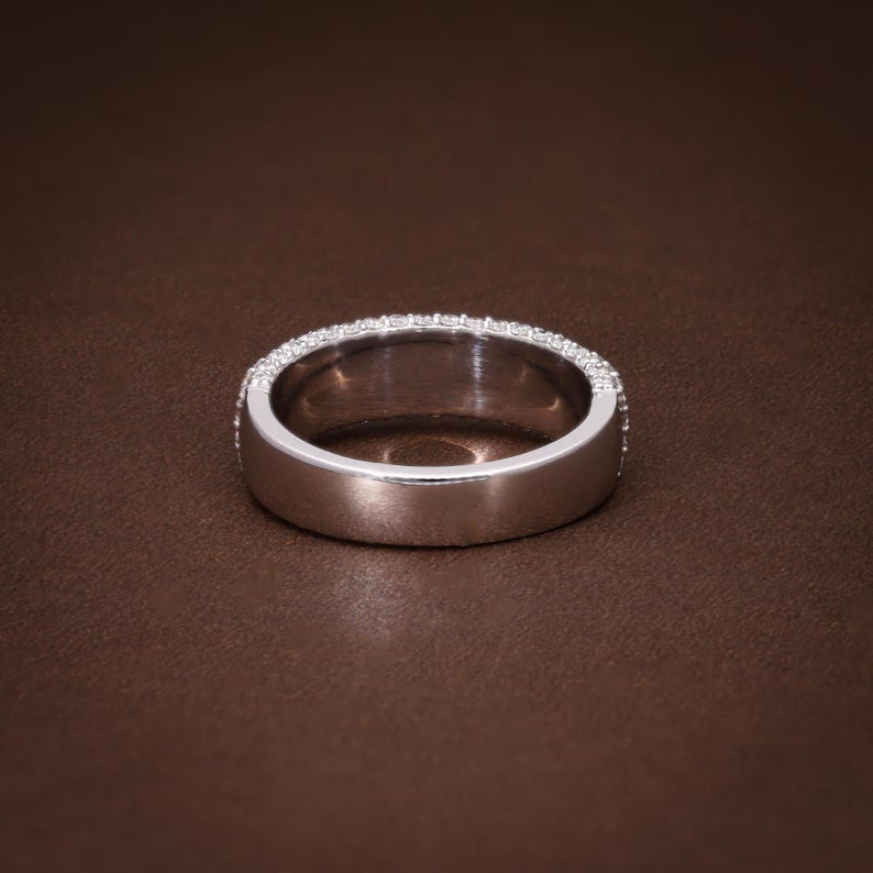 May include: A silver ring with a band of small, clear gemstones set around the top edge. The ring is set against a brown background, showcasing its polished metal and sparkling stones. A classic piece of jewelry.