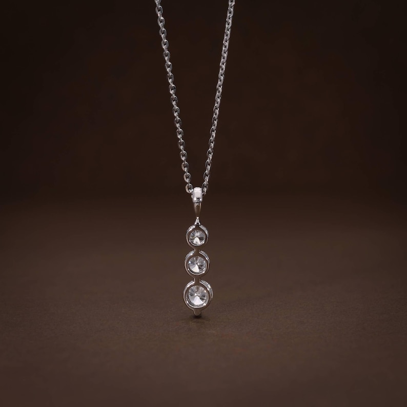May include: A silver pendant necklace featuring three clear, round gemstones of graduated sizes. The gemstones are set in individual bezels and suspended from a delicate silver chain. The necklace is displayed against a brown background.