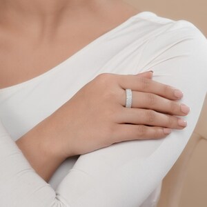 May include: A silver ring with multiple rows of small, clear stones. The ring is worn on a person's finger, and the hand is resting on a white long-sleeved shirt. The ring sparkles in the light.