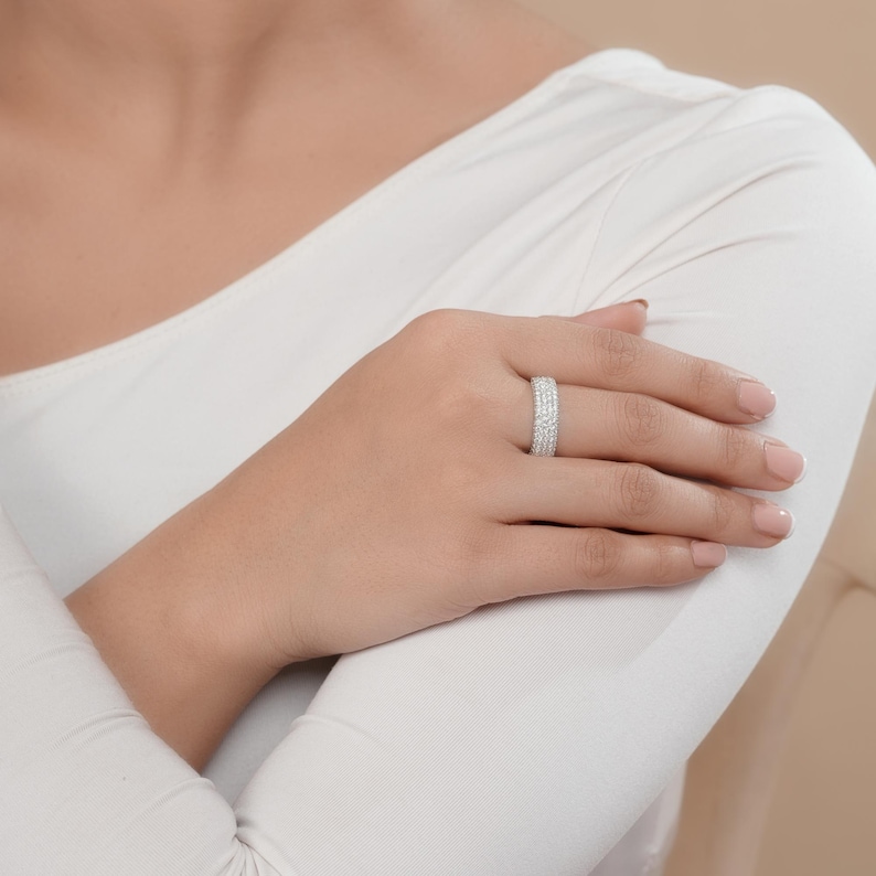May include: A silver ring with multiple rows of small, clear stones. The ring is worn on a person's finger, and the hand is resting on a white long-sleeved shirt. The ring sparkles in the light.