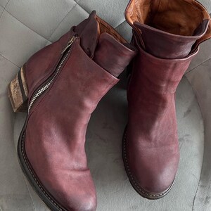 May include: A pair of ankle boots in a deep burgundy leather with a matte finish. The boots feature a side zipper closure, a rounded toe, and a stacked wooden heel with a dark brown sole. The interior lining is a contrasting tan leather.