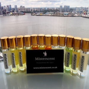 May include: A collection of 14 small, clear glass roll-on bottles with gold caps. The bottles are filled with a light yellow liquid. The bottles are arranged in a row on a white surface. A white card with the text "Misterscent" and the website "www.misterscent.co.uk" is in front of the bottles.