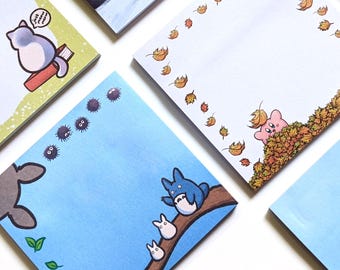 以下が含まれることがあります： 6つの正方形の付箋メモ帳。冬の帽子をかぶったキャラクターが描かれた星空、眠っているキャラクターが描かれた青いメモ帳、猫と本が描かれた緑のメモ帳、秋の葉が描かれた白いメモ帳、森の精霊が描かれた青いメモ帳、キノコとカエルが描かれた青いメモ帳のセット。