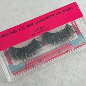 May include: A clear plastic case with a pink lid containing a pair of black mink eyelashes. The lid has the text "1-MINK LASHES 2-22-25MM 3-CRUELTY FREE 4-BLACK BAND" and the website address "www.presstillit.com".