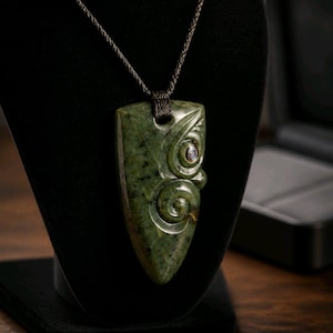 Op de afbeelding: Een schildvormige hanger van groene jade met uitgesneden spiraalpatronen en een kleine iriserende inleg. De hanger hangt aan een donkerbruin gevlochten koord en heeft een gepolijst oppervlak met natuurlijke donkere spikkels in de steen.
