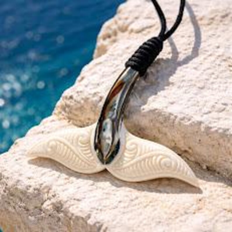 May include: A cream-colored, carved bone pendant in the shape of a whale tail, with a silver-colored metal accent. The pendant is suspended from a black cord. The background features a rocky surface and blue water.