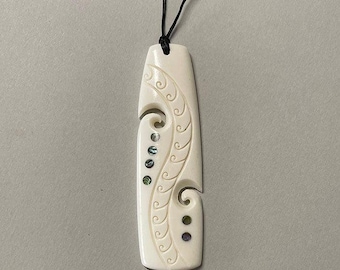 May include: A white bone pendant with a carved design and abalone shell accents. The pendant is attached to a black cord.