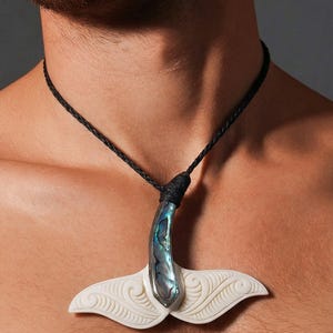 May include: A pendant necklace featuring a whale tail design. The pendant has a white, carved tail with a curved, iridescent abalone shell accent. The necklace has a black, braided cord.