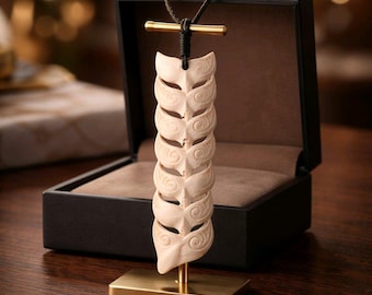 May include: A cream-colored carved pendant featuring a repeating spiral pattern, suspended from a dark cord on a gold-toned metal T-bar stand. The pendant is displayed in front of an open black box with a brown interior lining.
