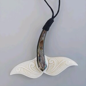 May include: A handcrafted pendant necklace featuring a whale tail design. The pendant has a white, carved tail with intricate patterns and a polished, iridescent abalone shell accent. The necklace has a black cord.