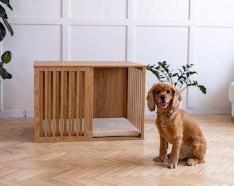 May include: A rectangular wooden dog crate with vertical slats on the front and side panels, featuring a light wood finish and a flat top surface. Inside the crate, a light-colored cushioned mat is visible on the floor of the enclosure.