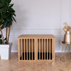 May include: A rectangular wooden pet crate featuring vertical slats on the front panel and a solid top surface. The crate is constructed from light-toned wood with a visible grain pattern and contains a dark black interior floor mat.