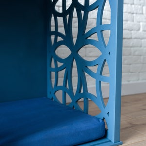 K&ouml;nnte beinhalten: Ein blaues, h&ouml;lzernes Haustierbett mit einem geometrischen Ausschnittmuster an der Vorderseite. Das Bett hat eine blaue Matratze im Inneren.