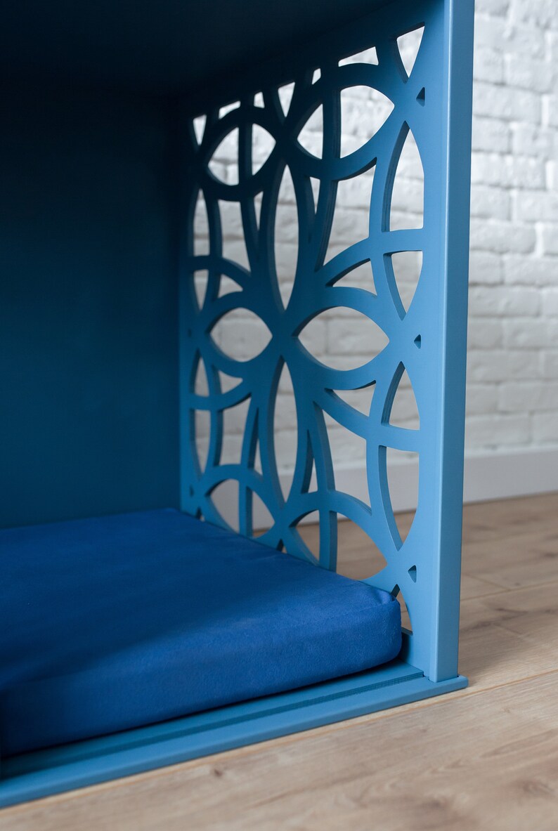 K&ouml;nnte beinhalten: Ein blaues, h&ouml;lzernes Haustierbett mit einem geometrischen Ausschnittmuster an der Vorderseite. Das Bett hat eine blaue Matratze im Inneren.