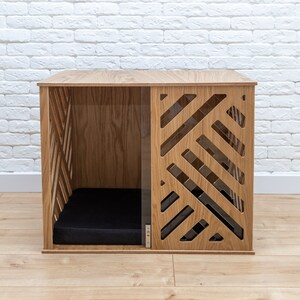 May include: A wooden dog crate with a geometric pattern on the front. The crate has a black cushion inside and is sitting on a wooden floor.