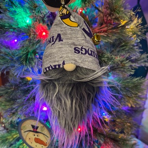 May include: A gray gnome ornament with a purple and yellow hat that says "Vikings" on it. The gnome is hanging on a Christmas tree with colorful lights.