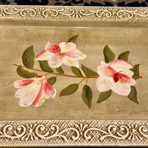 May include: A decorative ceramic serving tray with a raised, ornate border. The tray features a hand-painted design of pink and white flowers with green leaves on a light brown background.