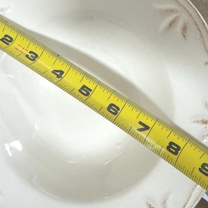 May include: A white ceramic bowl with a decorative rim and a yellow measuring tape across the top. The tape measures 9 inches.