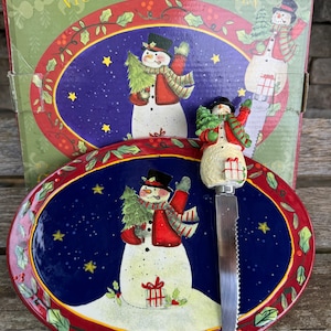 May include: A blue and red oval ceramic serving plate with a snowman design. The plate features a snowman holding a small Christmas tree and a red gift box. The plate comes with a matching snowman-shaped cheese knife.