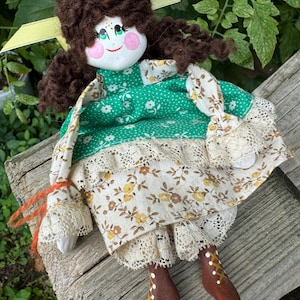 May include: A handmade doll with brown yarn hair, a green and white floral dress, and brown painted wooden legs and feet.