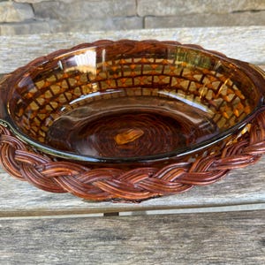 Puede incluir: Un plato para hornear de vidrio marrón con una base de mimbre tejido. El plato tiene un interior de vidrio transparente y un exterior marrón estampado.