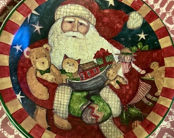 以下が含まれることがあります： サンタクロースが玩具に囲まれている絵が描かれた、クリスマスの装飾用プレート。 プレートには赤と金のストライプの縁と緑の縁があります。 イラストには、テディベア、猫、その他のお祝いの要素が含まれています。